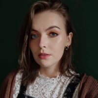 Portret młodej kobiety z półdługimi, lekko falowanymi włosami i jasną skórą. Ma na sobie bluzkę z koronkowym wykończeniem i ciemnymi detalami, a także brązowy sweter. Tło jest w ciemnozielonym kolorze, co kontrastuje z jej jasnymi oczami i makijażem.