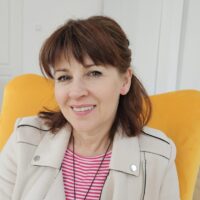 Kobieta z brązowymi włosami, uśmiechająca się, siedzi w żółtym fotelu, ubrana w jasną kurtkę i bluzkę w paski; w tle widać jasne pomieszczenie z drzwiami i białą szafą.