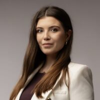 Kobieta w białym żakiecie z długimi, prostymi włosami, stojąca z założonymi rękami, na tle jednolitej szarej przestrzeni.