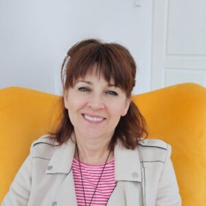 Kobieta z brązowymi włosami, uśmiechająca się, siedzi w żółtym fotelu, ubrana w jasną kurtkę i bluzkę w paski; w tle widać jasne pomieszczenie z drzwiami i białą szafą.